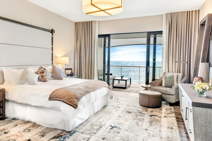 Modern Contemporary Interior Design by Fleur-de-lis Interior Design.This bedroom with a view of the ocean has neutral walls, a white bed frame, a grey armchair, a brown stool, and a rug in neutral.